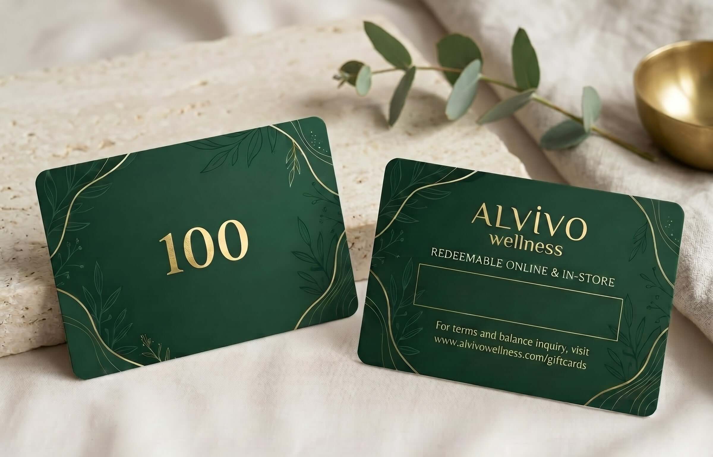 Two dark green gift cards with gold detailing are displayed on a soft beige fabric. One card shows the number 100 in gold, and the other shows the brand name 'ALYIVO wellness' with redemption text.